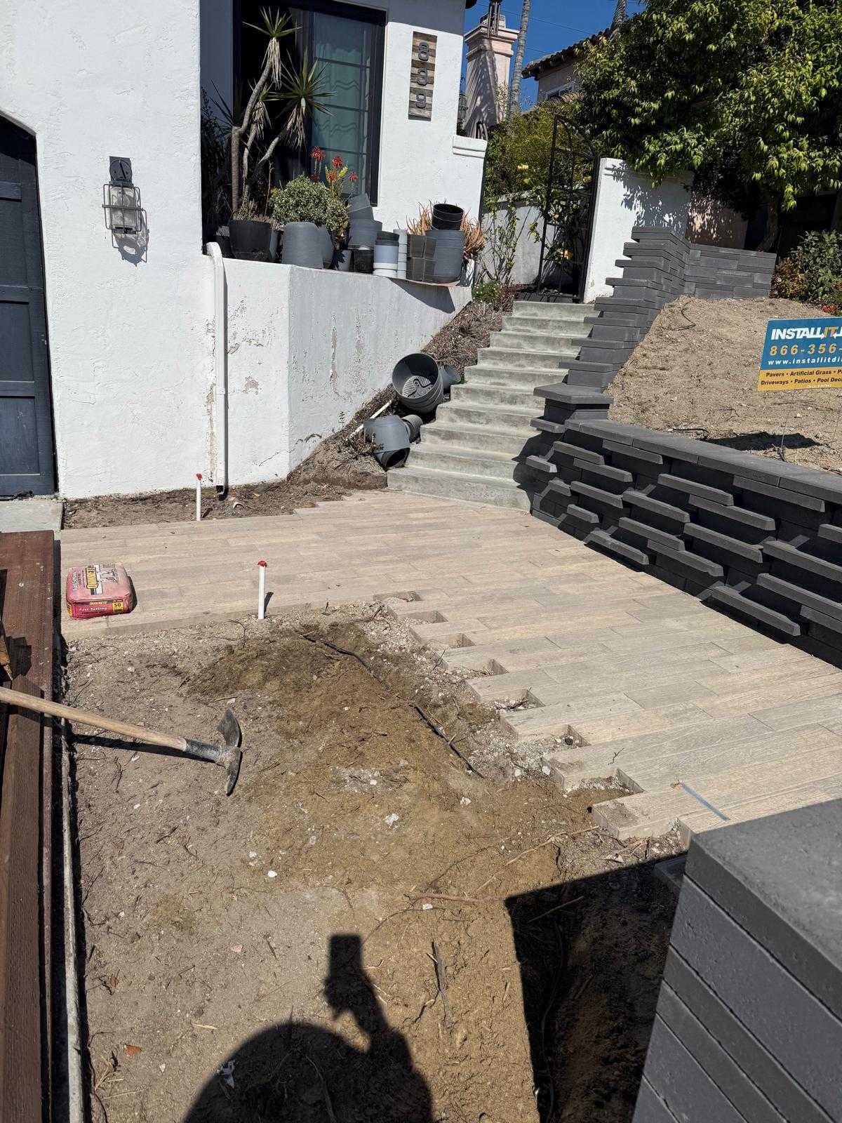 Partially installed paver driveway with stone retaining wall and tiled steps mid-construction, dirt still exposed at edges.