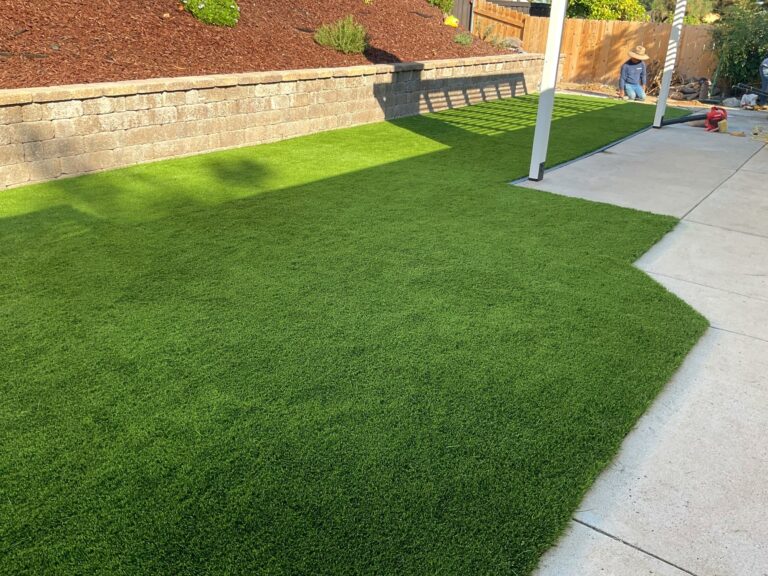 Freshly installed synthetic turf lawn beside a concrete patio, with a two-tier segmental retaining wall and red mulch slop...