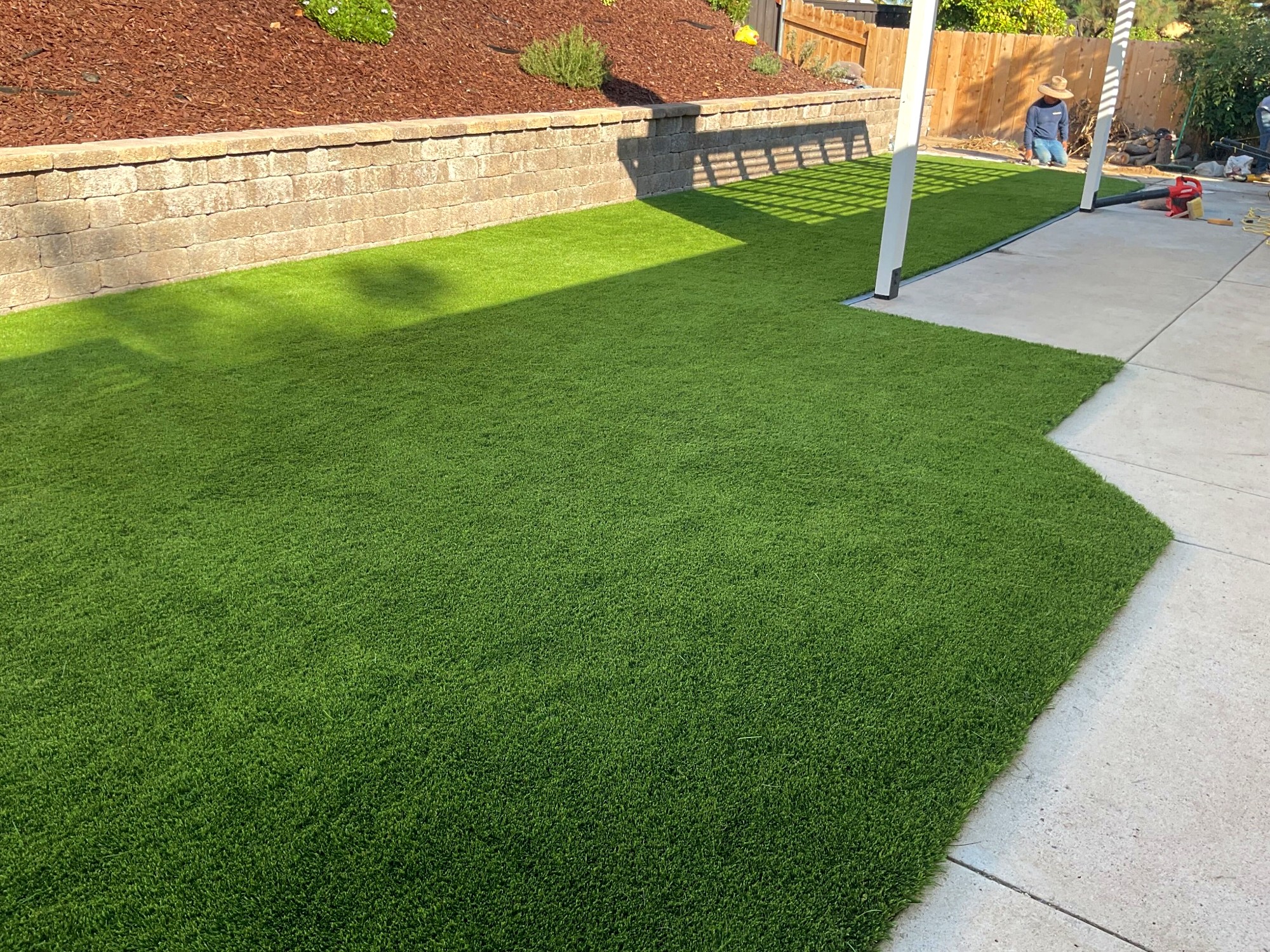 Synthetic turf lawn running the length of a concrete patio, bordered by a capped segmental retaining wall and a red-mulch slope.