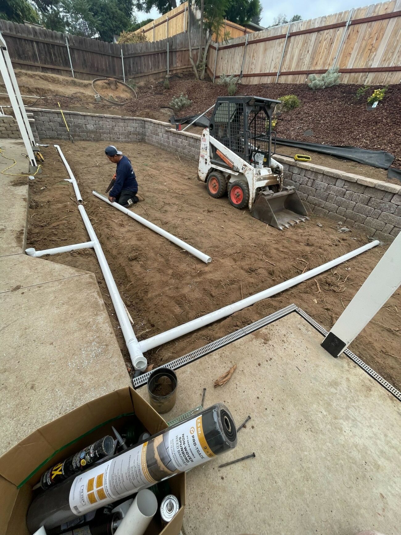 Worker laying white PVC drainage pipe across graded dirt in a walled backyard, with a Bobcat skid steer nearby.