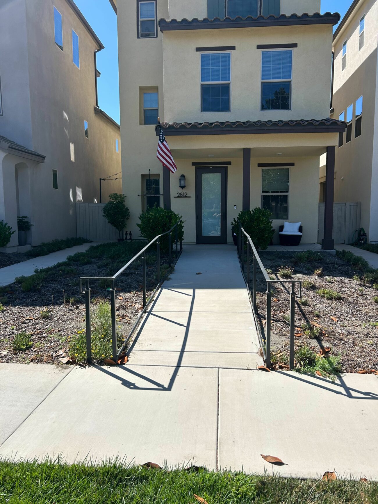 Concrete front walkway with metal handrails leading to a residential front entry and covered porch with columns. Landscaped planting beds with mulch and drought-tolerant plants flank the walkway and a narrow grass strip is visible at the sidewalk edge.