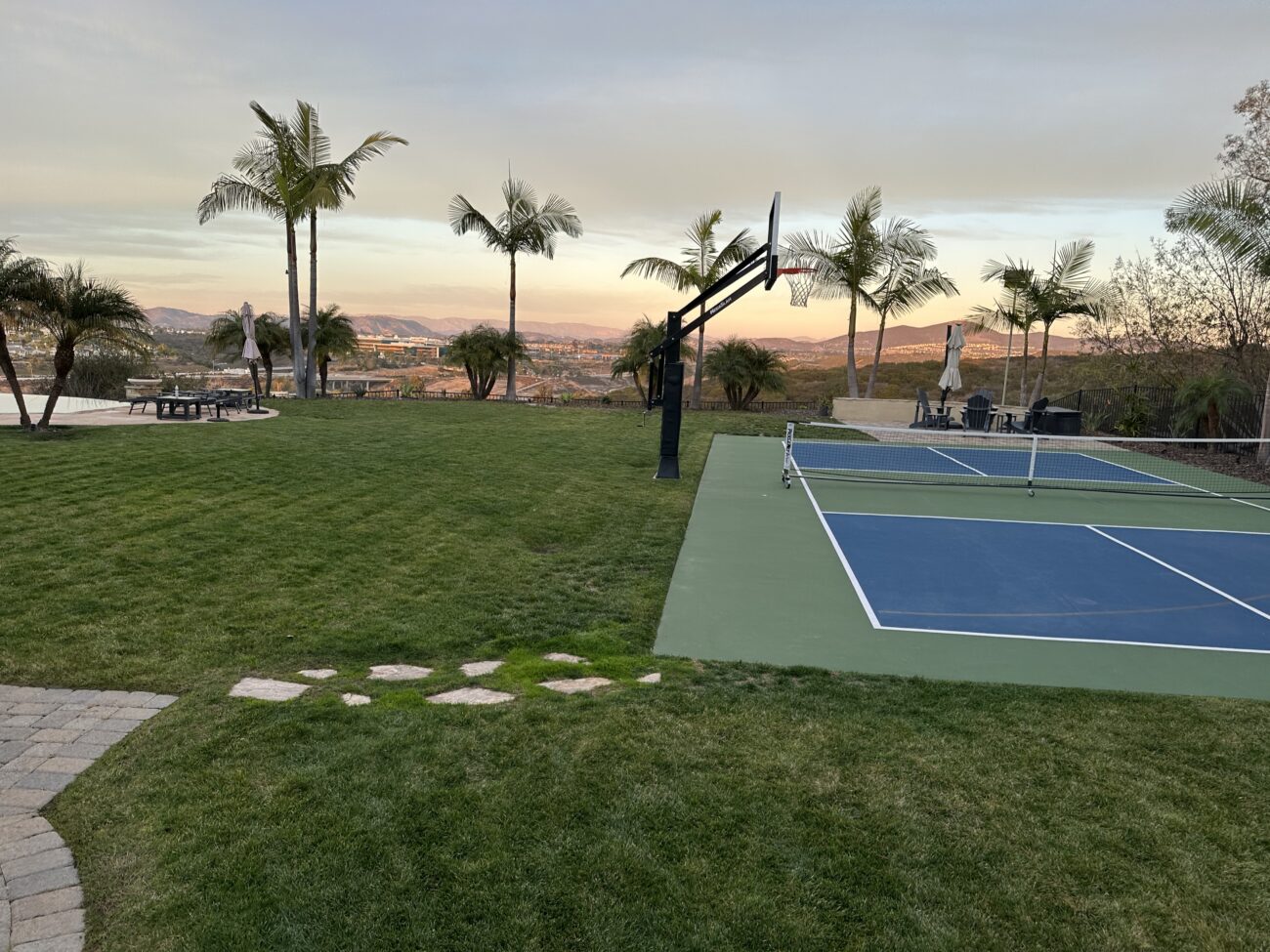 Residential backyard showing a completed landscape with a planted lawn and interlocking paver walkway adjacent to a painted sports court and pool deck. The scene includes natural turf, stepping stones, palm trees and finished hardscape elements indicating final installation work.