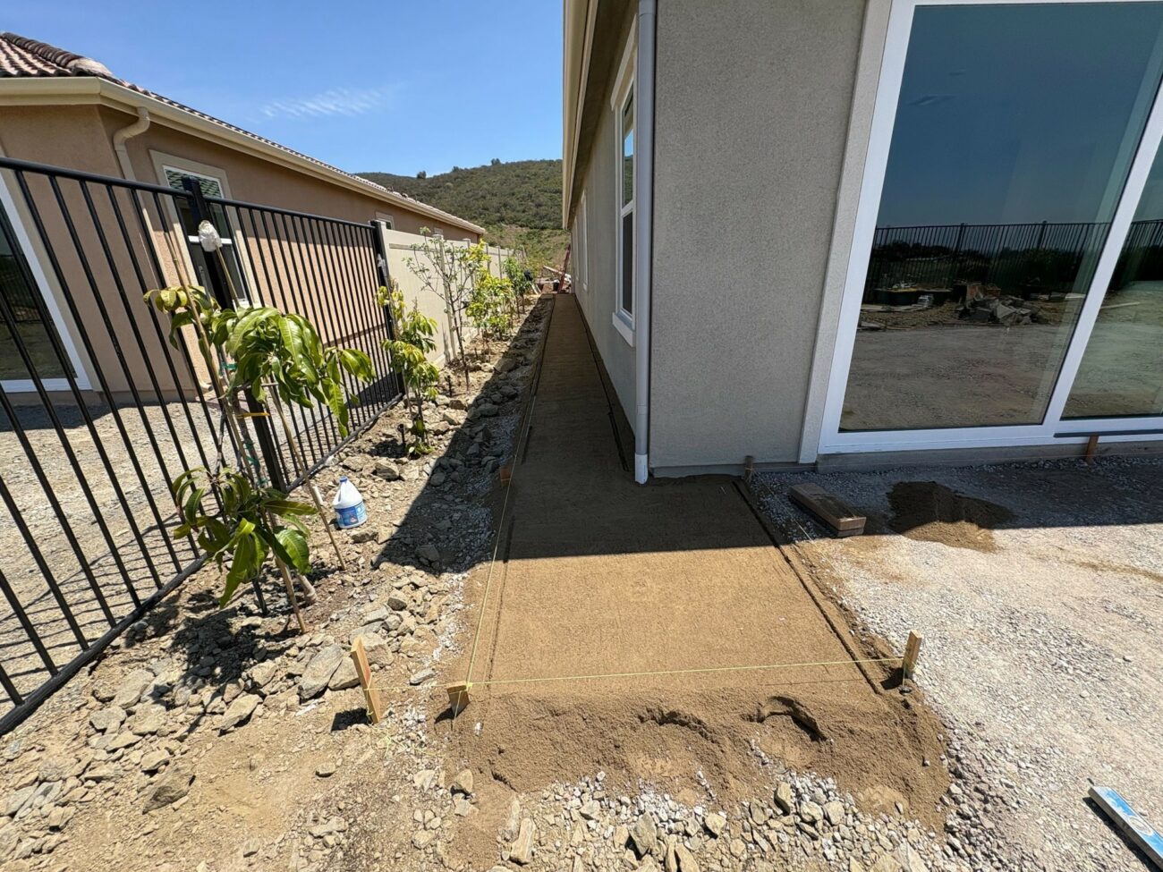 Freshly compacted sand base laid along a home's side yard, with wooden stakes and string lines guiding paver walkway layout.