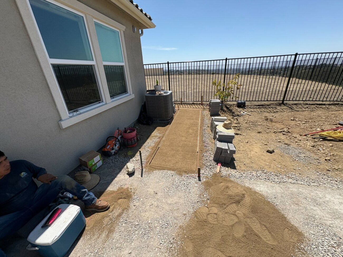 Worker resting against stucco house wall beside sand base and wood forms prepped for paver walkway installation, with stac...
