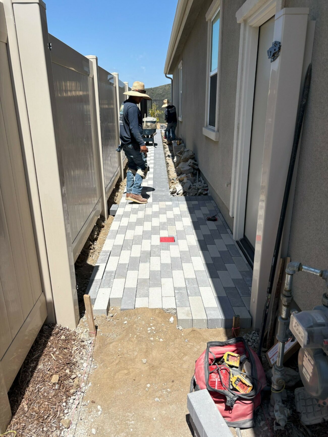 Two workers installing a two-tone gray interlocking paver walkway in a narrow side yard between a house and vinyl fence.