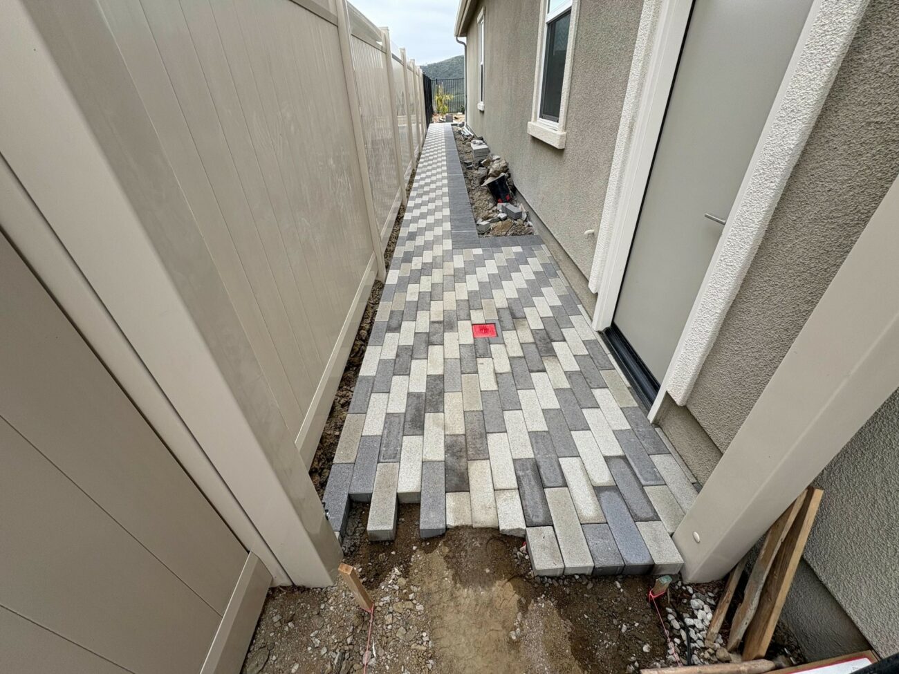 Narrow side-yard walkway with gray and white interlocking pavers laid in a running bond pattern between a stucco house and...