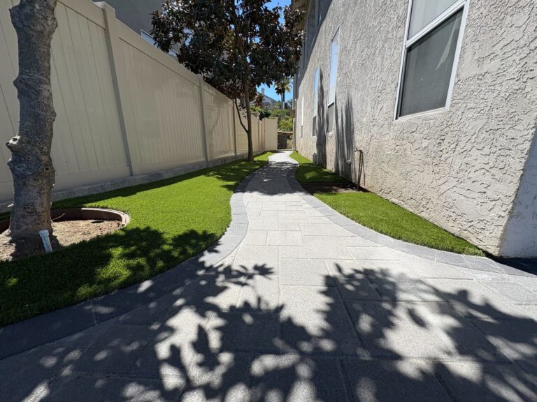 Winding stone paver walkway flanked by bright artificial turf between a stucco building and a white vinyl fence.