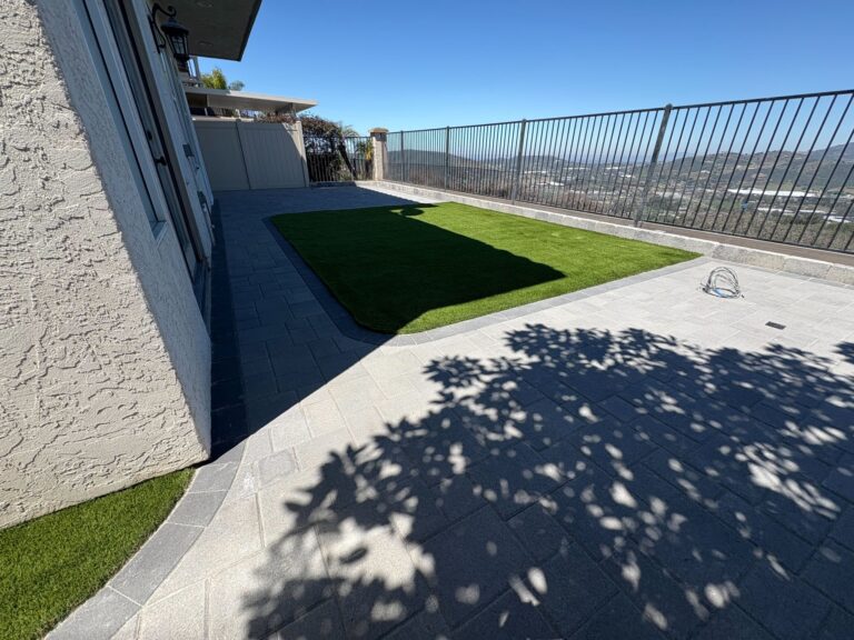 Backyard patio with dark charcoal pavers, a rectangular artificial turf section, and wrought-iron railing overlooking a va...