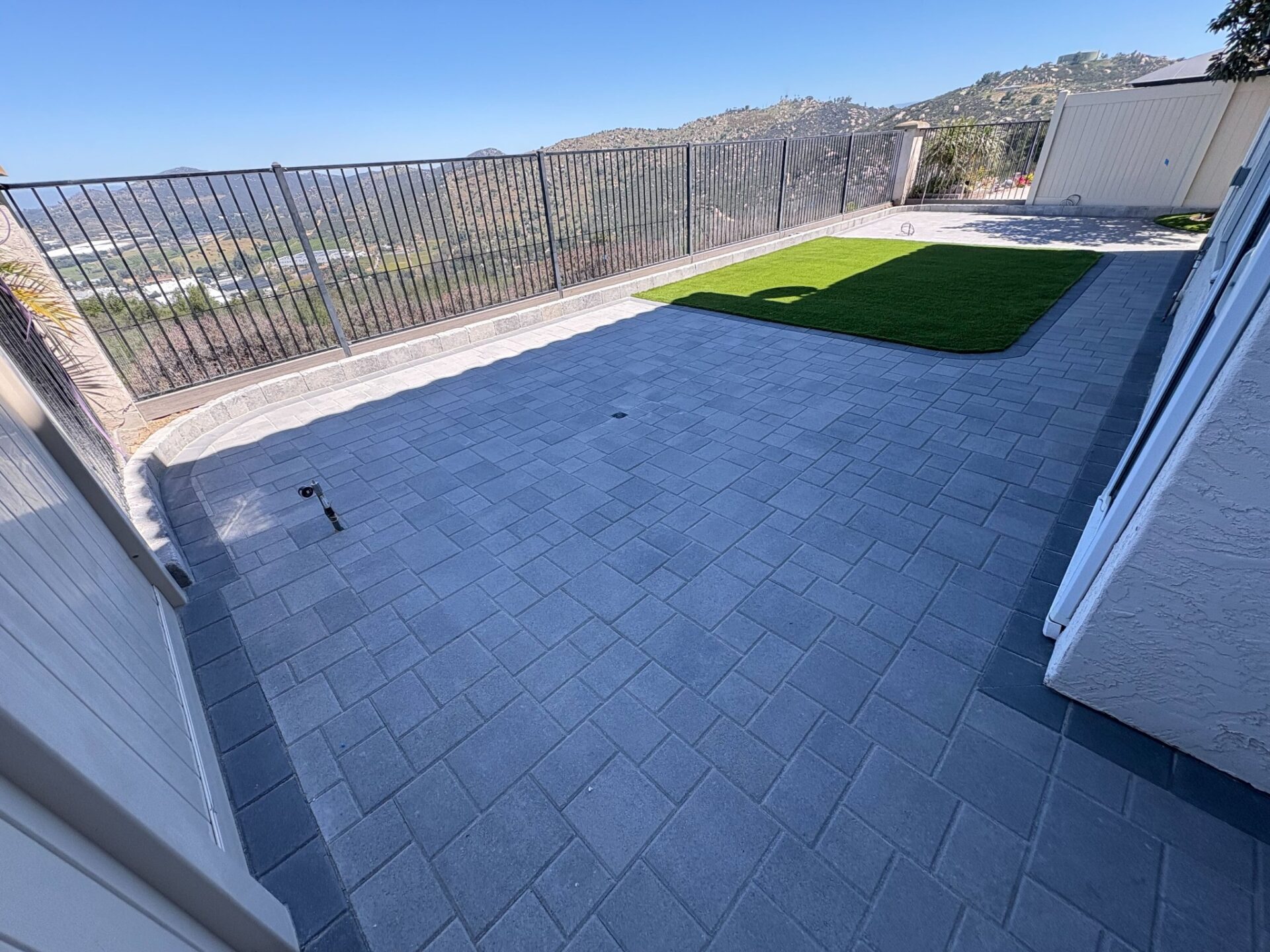 Gray paver patio with a small artificial turf section and wrought-iron railing overlooking a valley and rocky hills in Esc...