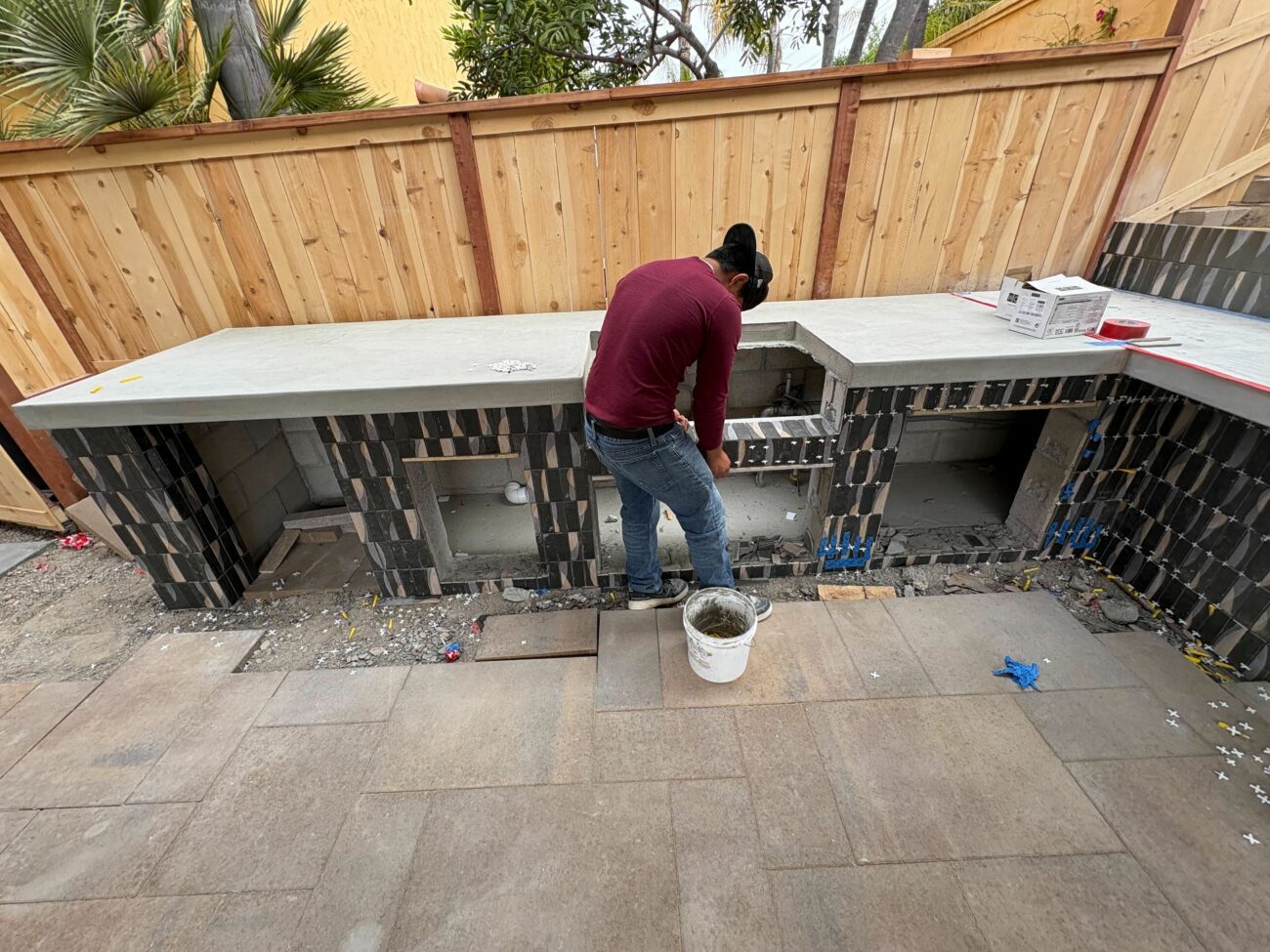 Tile setter applying dark stone veneer to an L-shaped outdoor kitchen frame with a poured concrete countertop in progress.