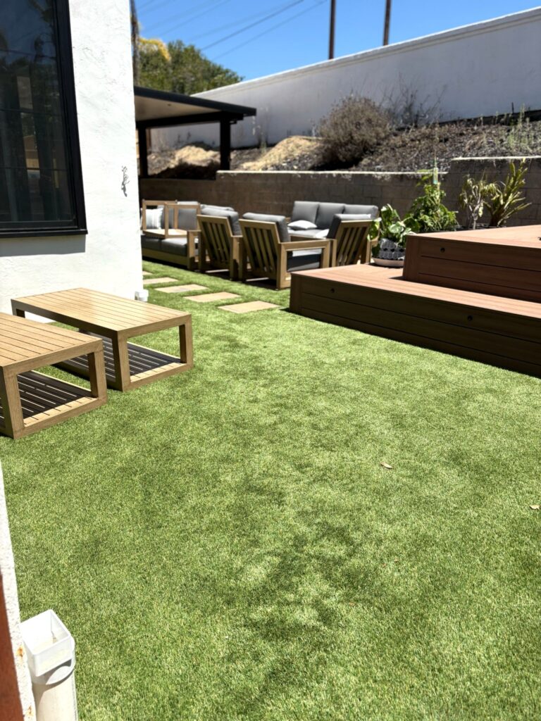Artificial turf lawn flanked by teak benches and a raised composite deck with outdoor seating in the background.