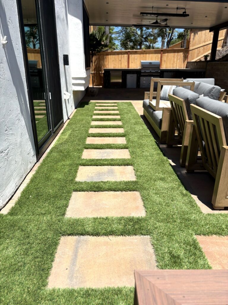 Belgard Moduline 24x24 stepping stones set in artificial turf lead toward a covered outdoor kitchen and lounge area.
