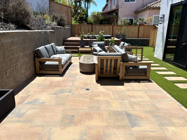 Teak outdoor sectional and armchairs围绕a concrete fire pit on tan pavers, with artificial turf and stepping stones alongside.