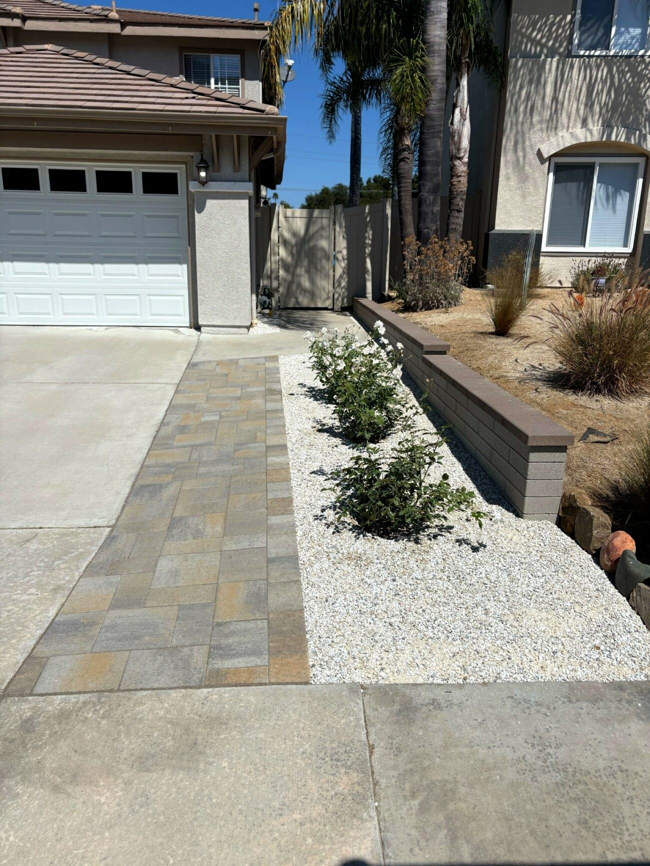 Finished front yard hardscape showing interlocking pavers forming a driveway/walkway edge adjacent to a concrete driveway, with a gravel planting strip containing small shrubs. A low segmental retaining/seat wall with stone cap separates the planted area from the raised yard and home exterior, indicating a completed landscape installation.