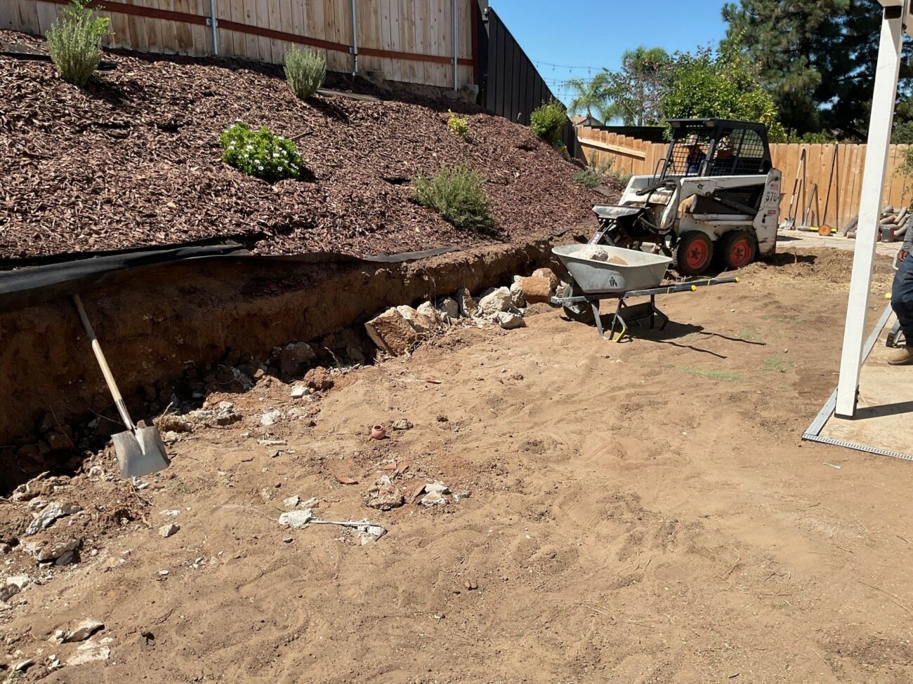 Excavated slope with mulch ground cover, a bobcat skid steer, wheelbarrow, and shovel during backyard demolition work.