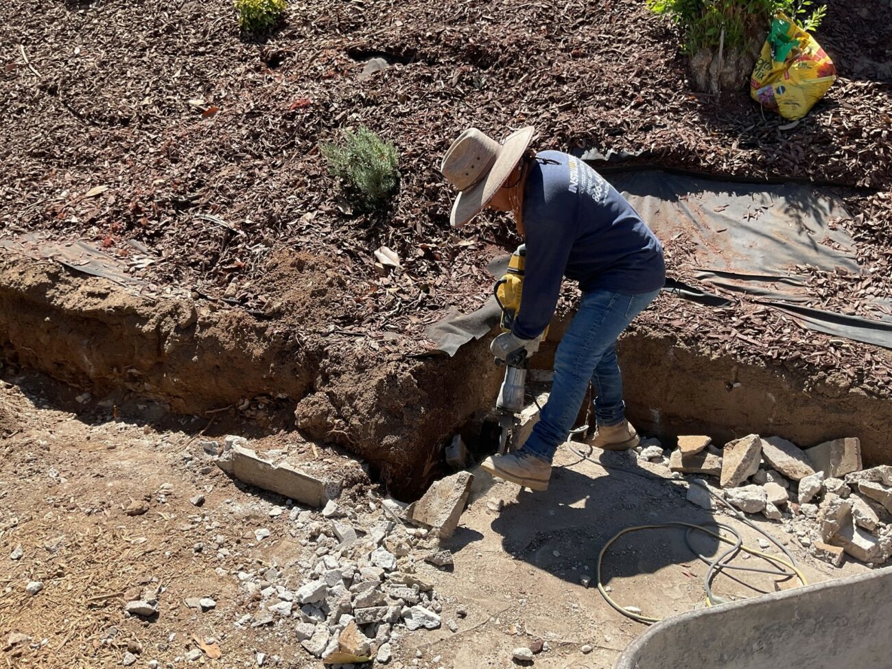 Worker using a pneumatic jackhammer to break up an existing concrete retaining wall edge during demolition.