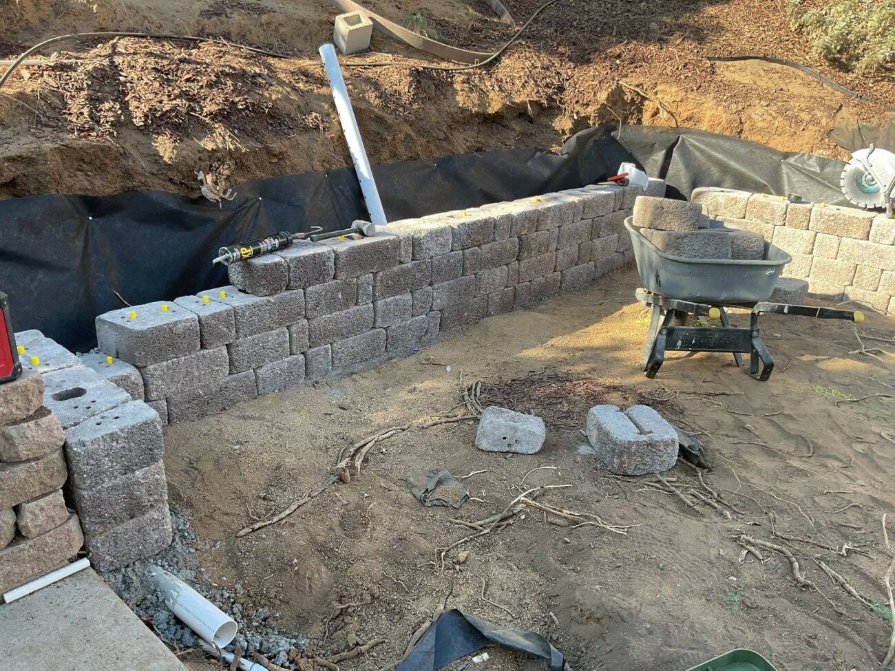 Partially built segmental retaining wall with stacked gray block units, a wheelbarrow with cap stones, and sandy excavated...