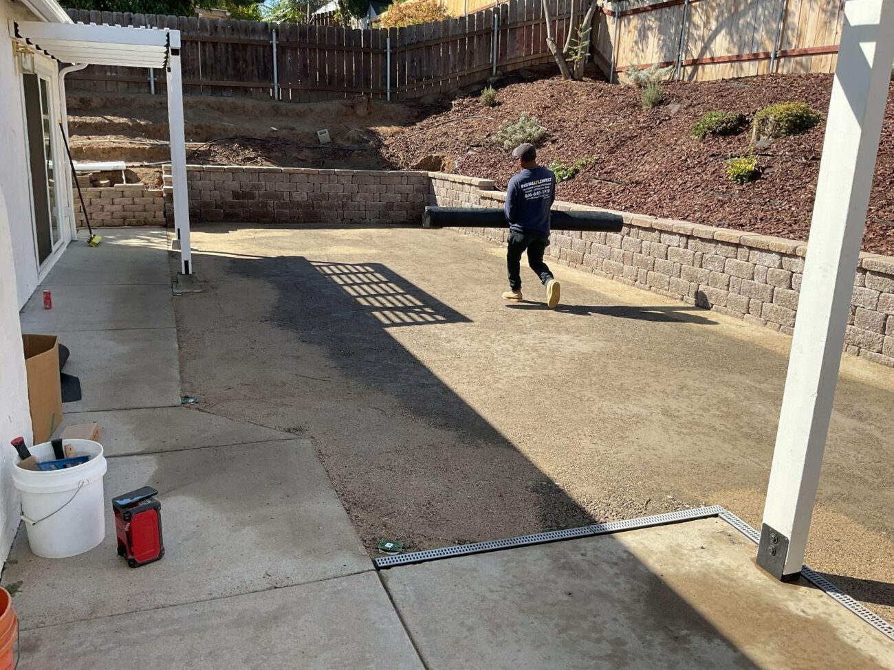 Worker carrying rolled synthetic turf across a freshly poured concrete patio with a completed segmental retaining wall beh...