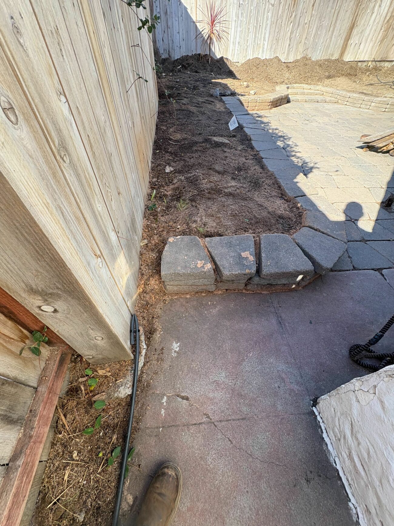 Narrow side-yard planting bed bordered by granite pavers alongside a wood fence, mid-construction.
