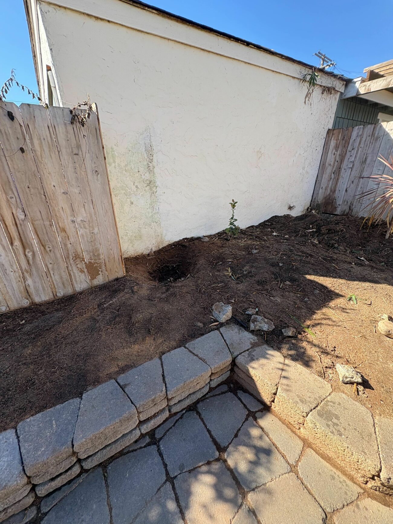 Raised planting bed with dark mulch and a low segmental retaining wall, bordered by a paver patio beside a stucco wall.