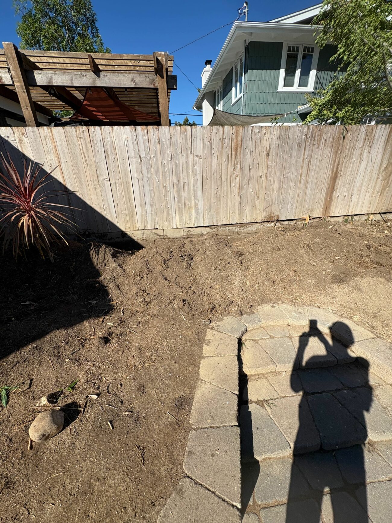 Bare dirt backyard cleared for construction, with an existing paver patio edge and a wooden pergola visible.