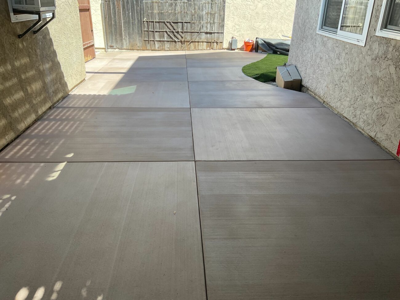Finished broom-finished concrete slab panels with visible control joints forming large squares, adjacent to a small artificial turf patch next to the house. The image shows a clean residential side-yard/patio area with a materials box near the wall, indicating a completed outdoor surface installation.