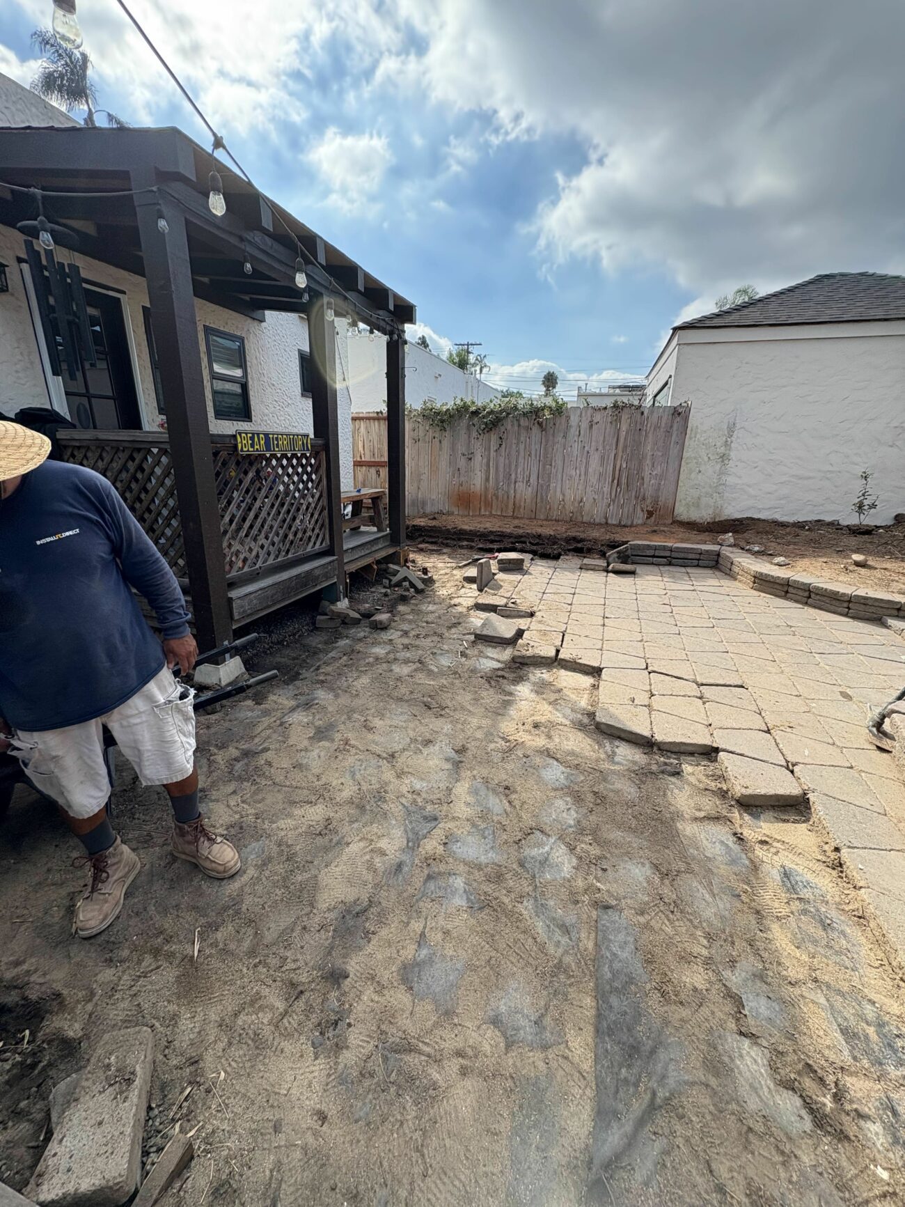 Worker in straw hat standing on compacted sand base beside a partially laid paver patio in a residential backyard.