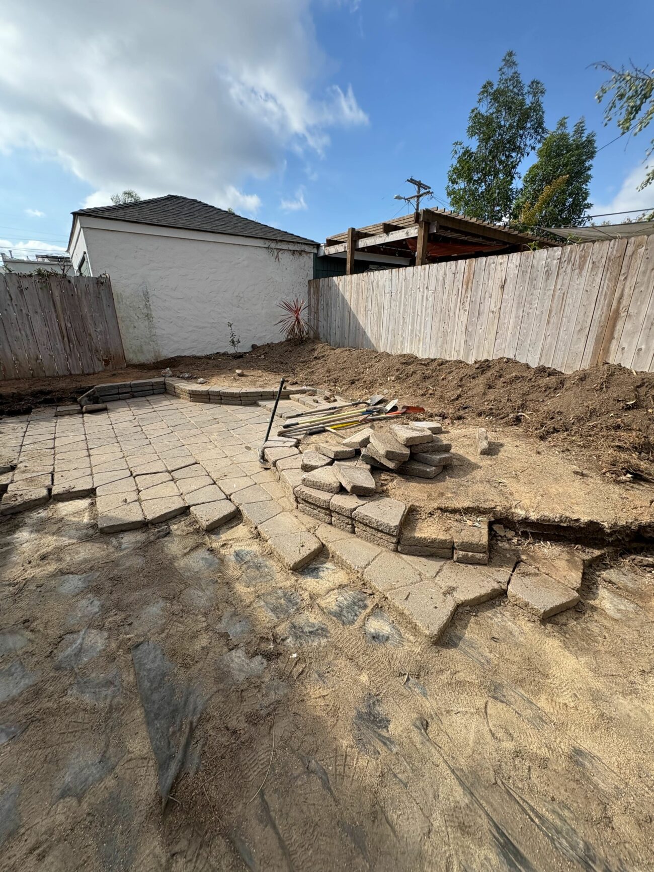 Partially laid concrete paver patio mid-installation in a fenced backyard, with stacked pavers and tools on bare soil.