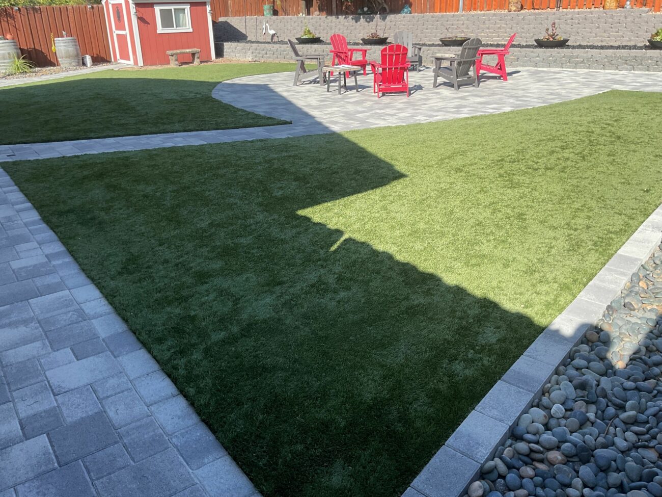 Finished backyard showing interlocking paver patio and walkways with artificial turf infill and a segmental retaining wall with planter beds. River rock edging and outdoor seating with red Adirondack chairs complete the landscaped hardscape.