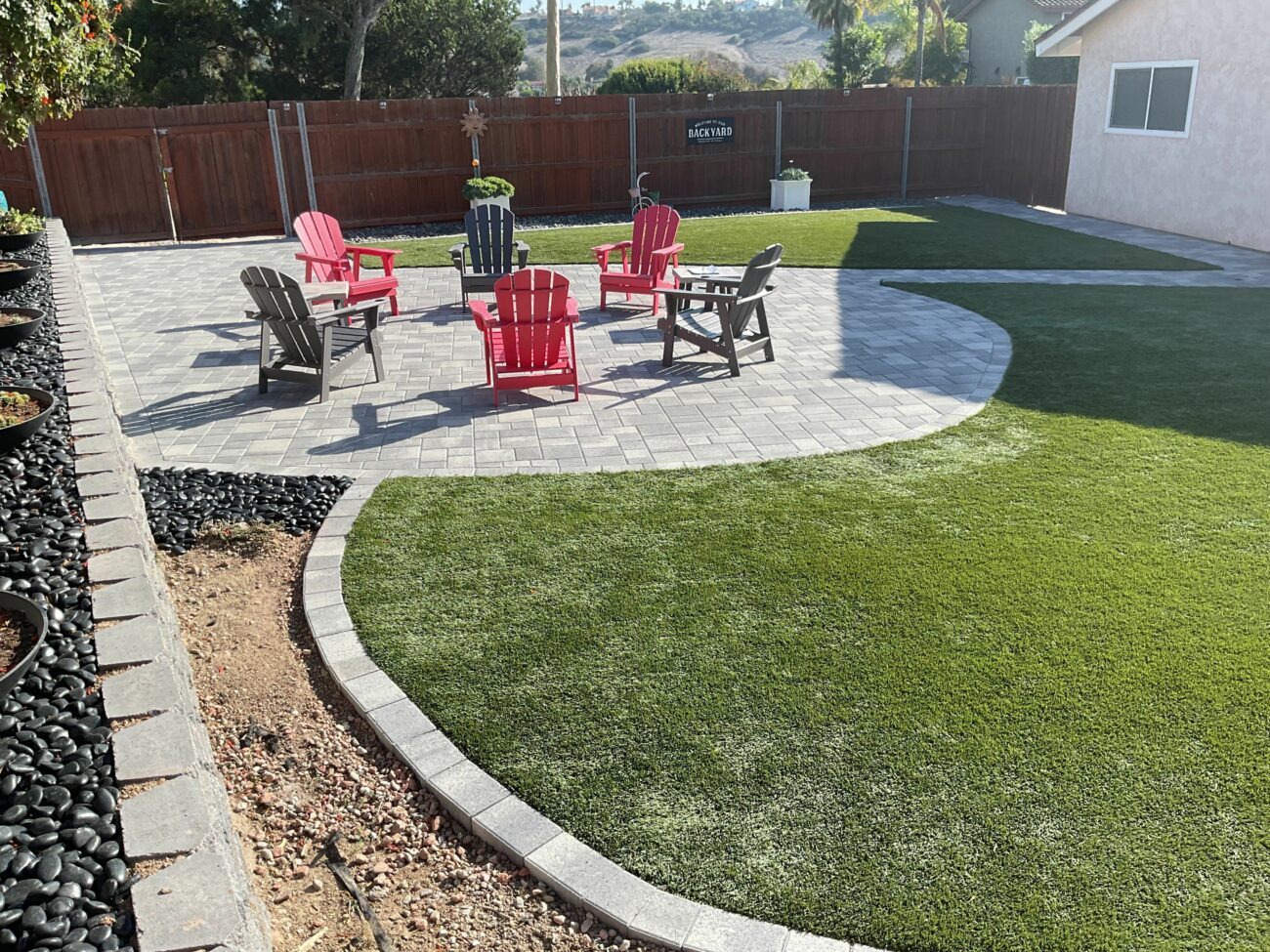 Finished residential backyard showing an interlocking paver patio with a curved edge abutting artificial turf and a segmental retaining wall with river rock and planter pockets. Outdoor seating is staged on the patio, indicating project closeout and a completed landscape installation.