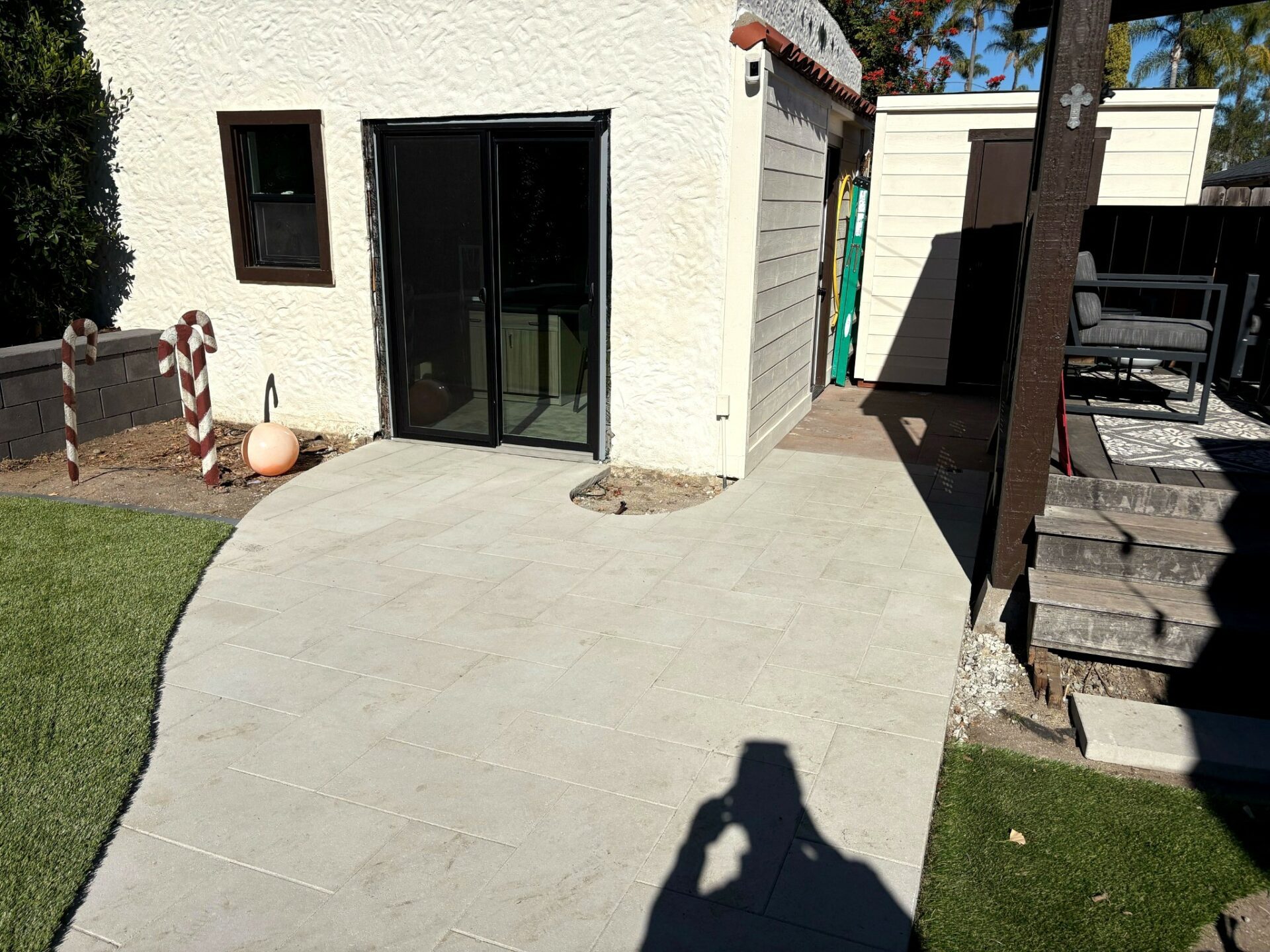 Light gray large-format paver patio and curved walkway beside a white stucco home with black sliding glass doors and artif...