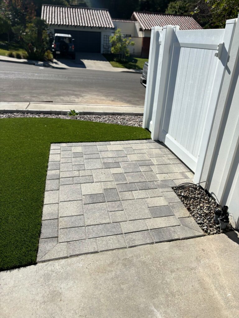 The interlocking-paver landing at the side gate, where the artificial turf meets the hardscape in a crisp, straight edge.