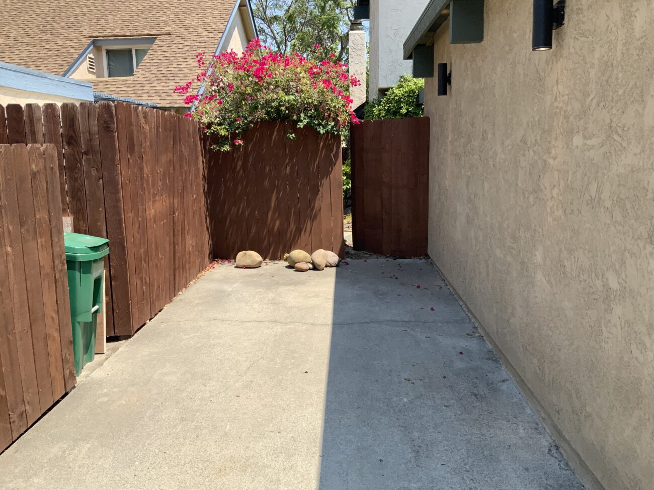Narrow side yard showing an existing concrete walkway/slab between a stucco house wall and a wood fence. A planter with flowering bougainvillea and several boulders sits at the far end; wall-mounted exterior lights are visible on the house. No construction activity or materials present, suitable for pre-construction assessment.