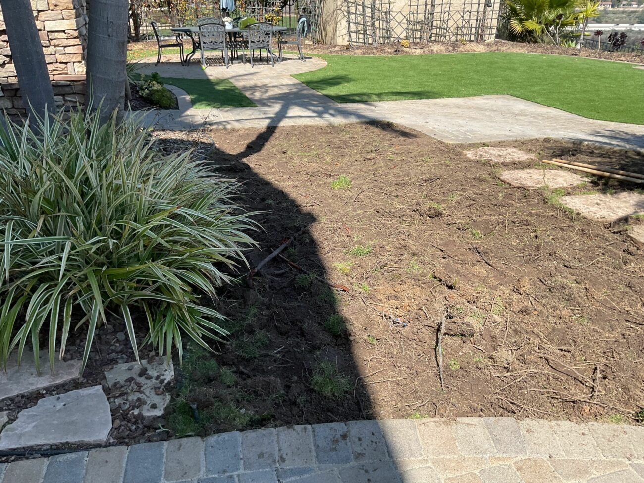 Backyard construction scene showing a paver walkway and patio in the foreground, with artificial turf installed in the background and an adjacent area of exposed soil where turf or planting has been removed. Patio furniture and a segmental retaining wall are visible; the site appears to be in mid-installation with base prep underway for additional turf or landscape work.