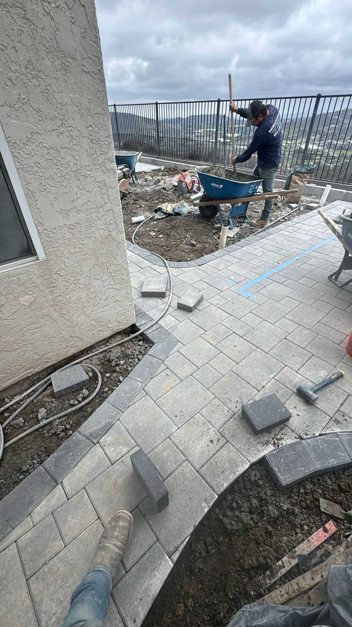 Worker shoveling material into a wheelbarrow on a partially laid gray paver patio with loose pavers and a rubber mallet ne...