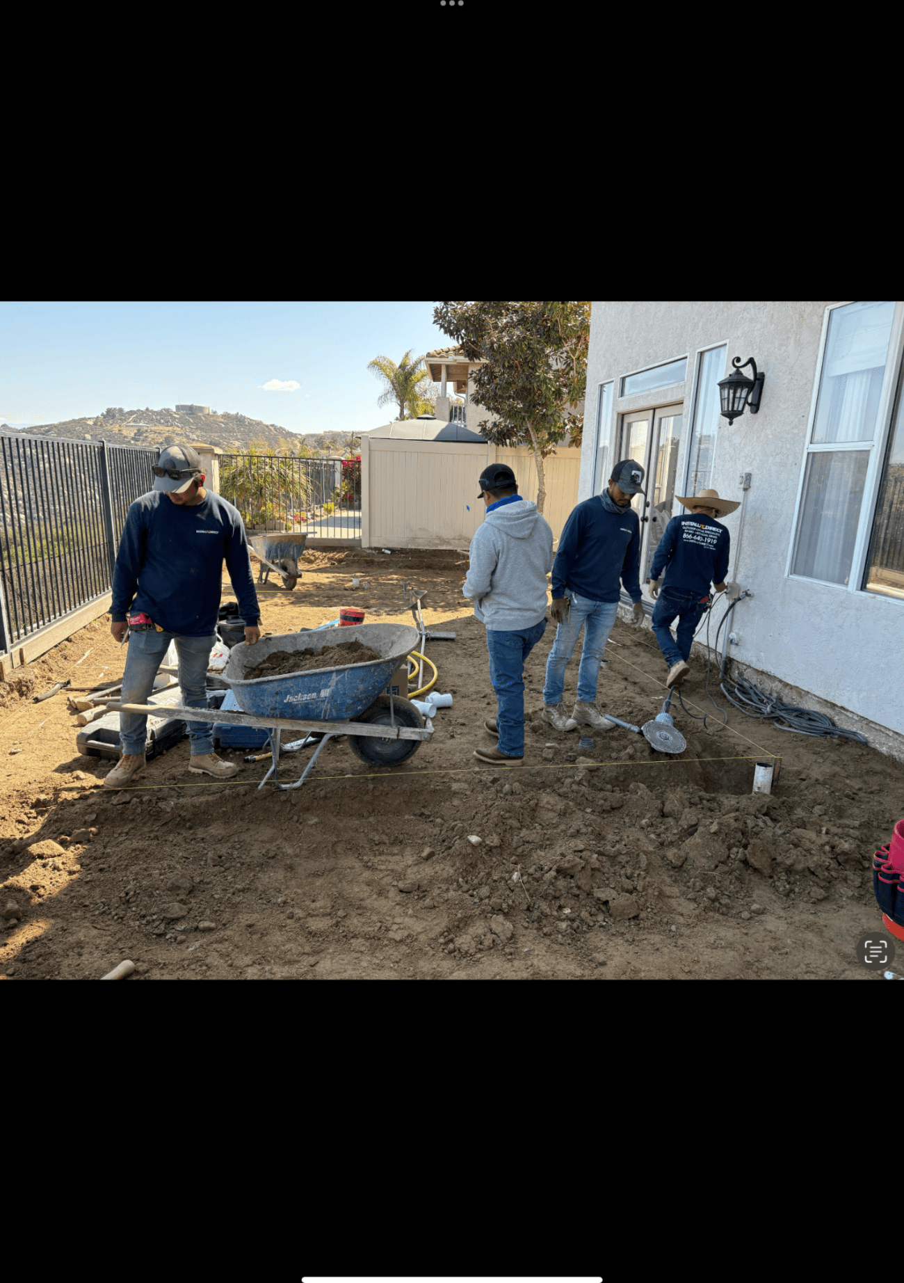 Four workers excavating a residential backyard, with a loaded wheelbarrow and string lines marking the layout on bare dirt.