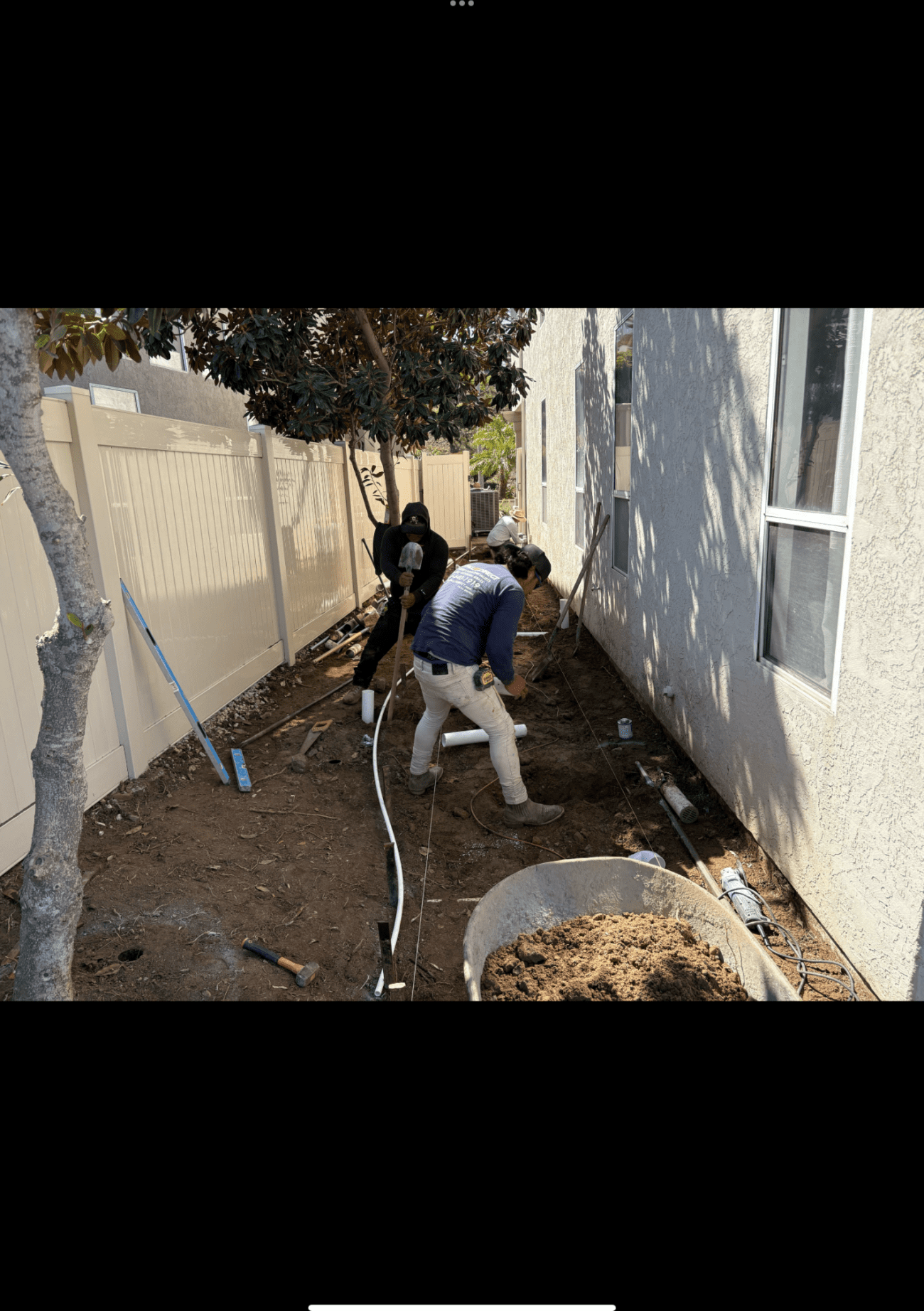 Two workers digging and laying PVC drainpipe in a narrow dirt side yard beside a stucco house.