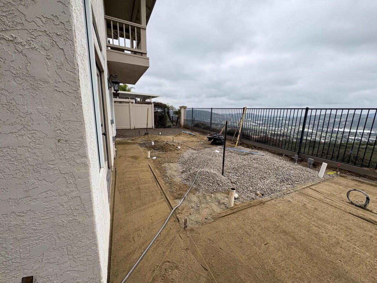 Backyard under construction with gravel base, conduit lines, and drain stubs on compacted soil beside a stucco home in Esc...