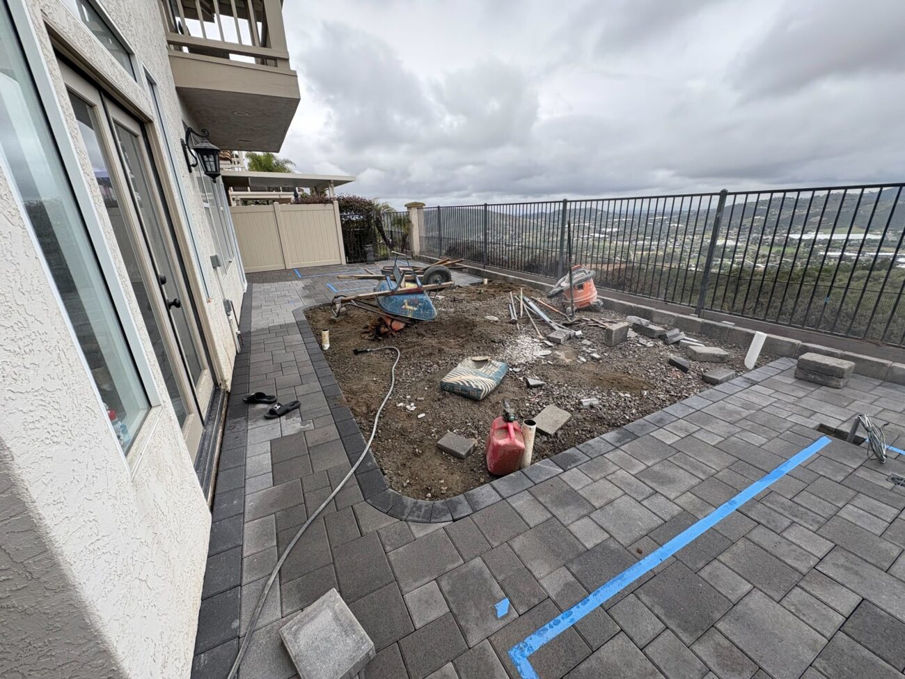 Paver patio partially installed around a dirt center section with a wheelbarrow, gas can, and construction tools on a hill...