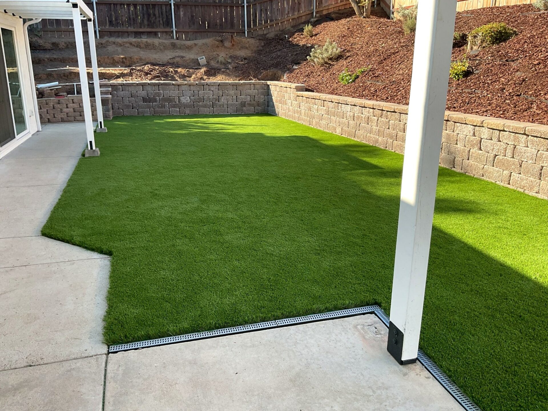 Finished turf lawn meets the patio at a channel drain, a segmental retaining wall stepping down the mulched slope behind it.