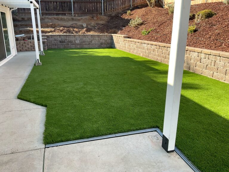 Freshly installed synthetic turf lawn bordered by a segmental block retaining wall, red mulch slopes, and a concrete patio...