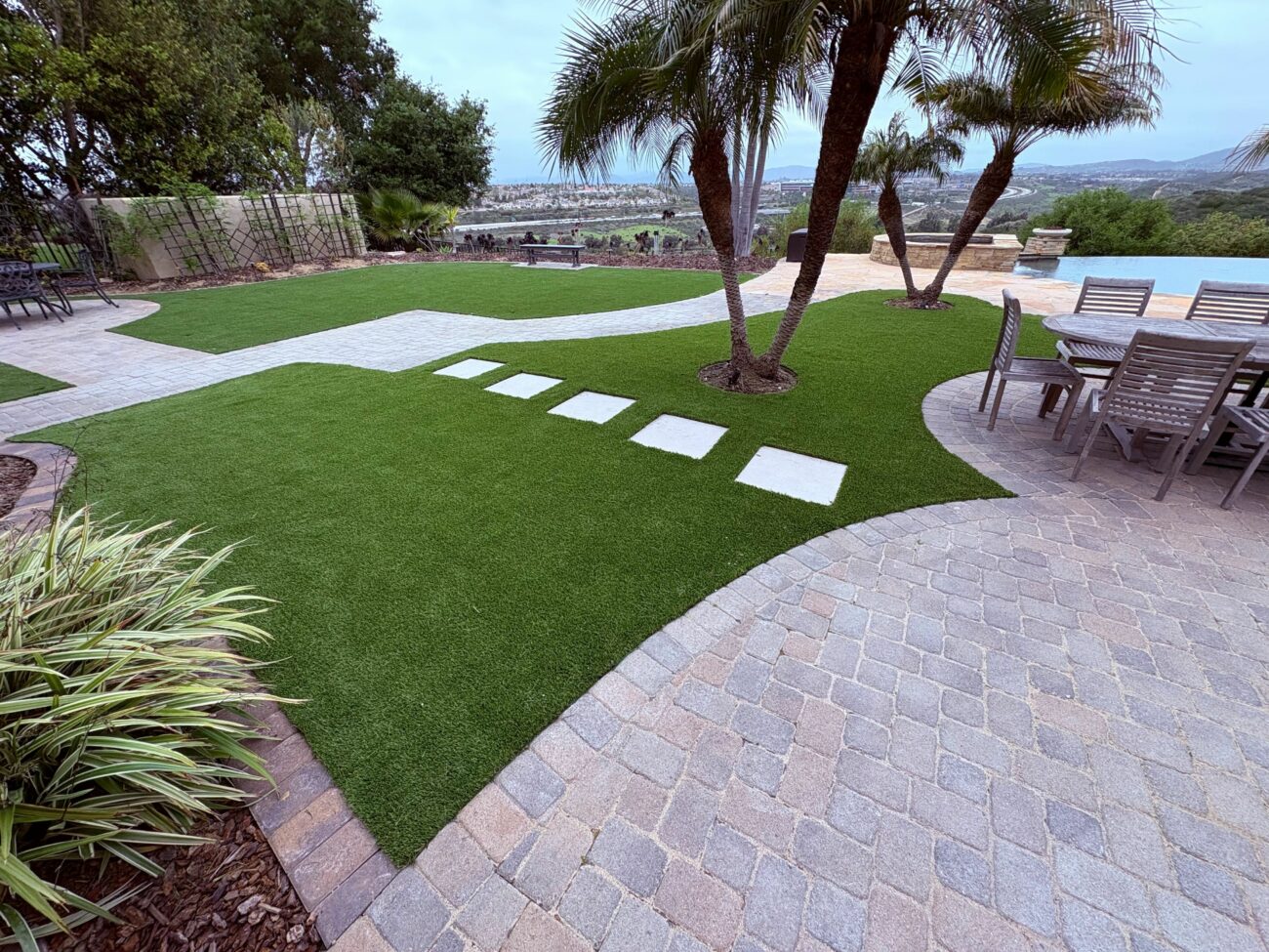 Finished backyard hardscape showing synthetic turf installation bordered by interlocking paver patio and pool deck. Concrete square stepping pads set into the turf create a walkway to a seating/dining area beneath palm trees and overlook a pool area and distant view.
