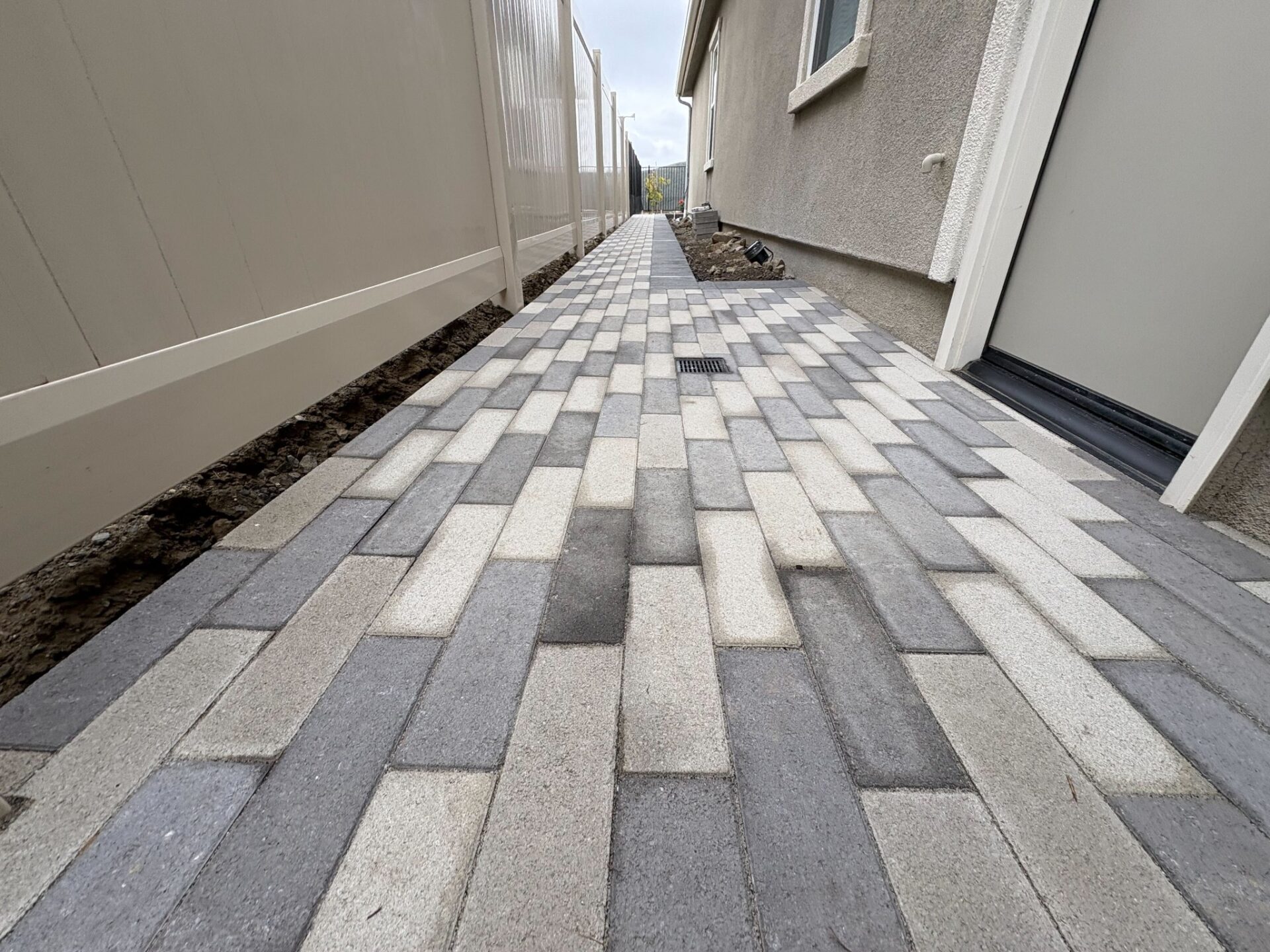 Narrow side-yard walkway paved with mixed light and dark gray interlocking pavers between a stucco house and a fence.