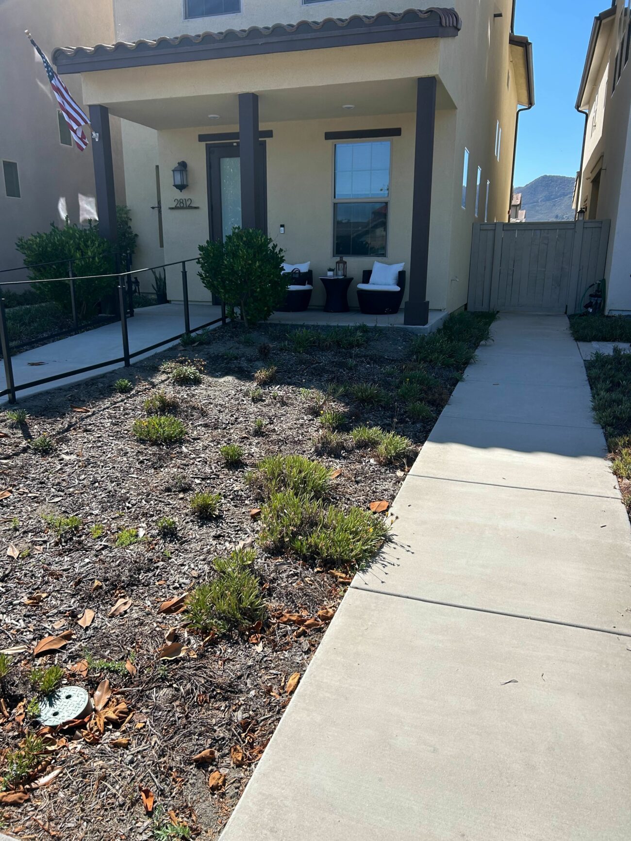 Front yard and entry porch showing a concrete walkway and ramp with railing leading to a covered porch. The planting bed beside the walkway is mulched with sparse drought-tolerant shrubs, presenting a clear area for pavers or artificial turf installation.