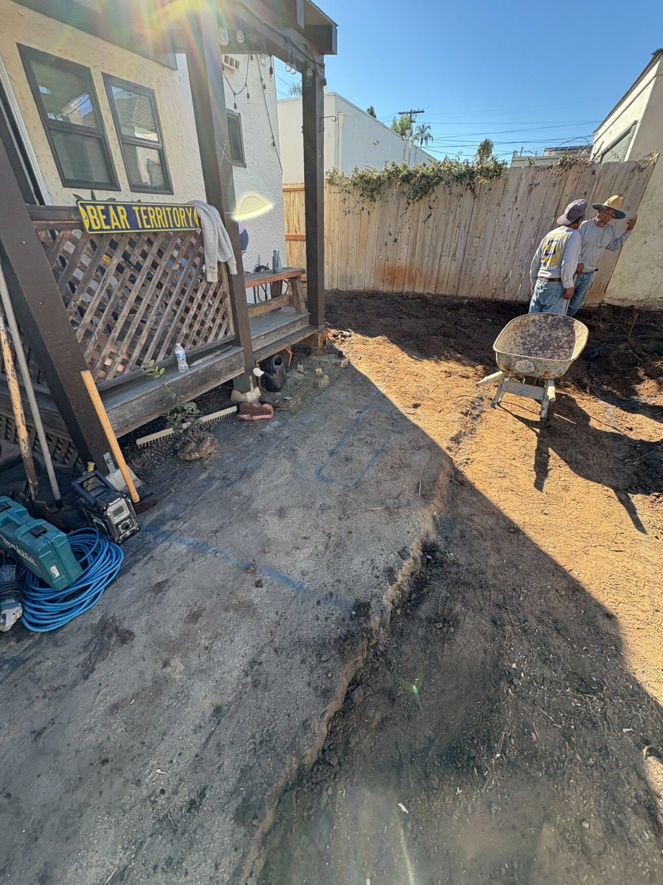 Two workers with a wheelbarrow clearing and grading a bare backyard in preparation for hardscape installation.