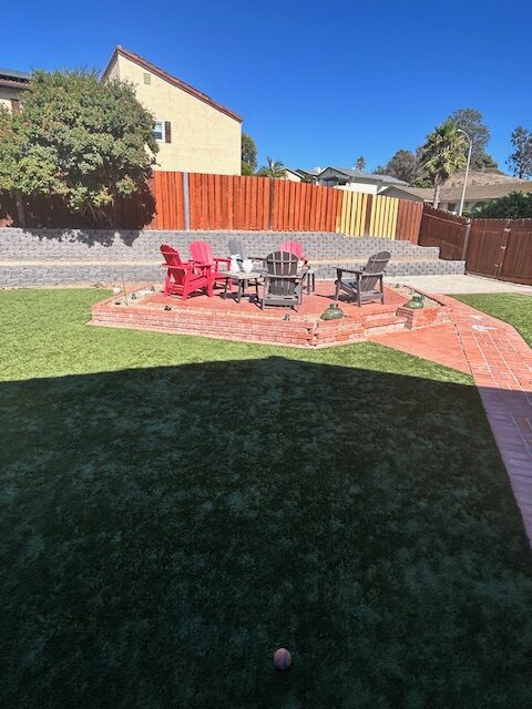 Finished backyard showing an interlocking paver patio with seating and a central fire pit, bordered by artificial turf. A segmental retaining wall and newly stained wood fence run along the rear property line with the house exterior visible beyond.