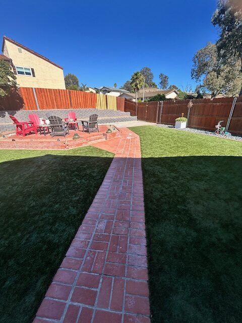 Finished residential backyard showing a straight brick paver walkway leading to a raised paver patio with seating. Artificial turf has been installed on both sides of the walkway and a concrete block retaining wall and new wood fence define the perimeter.