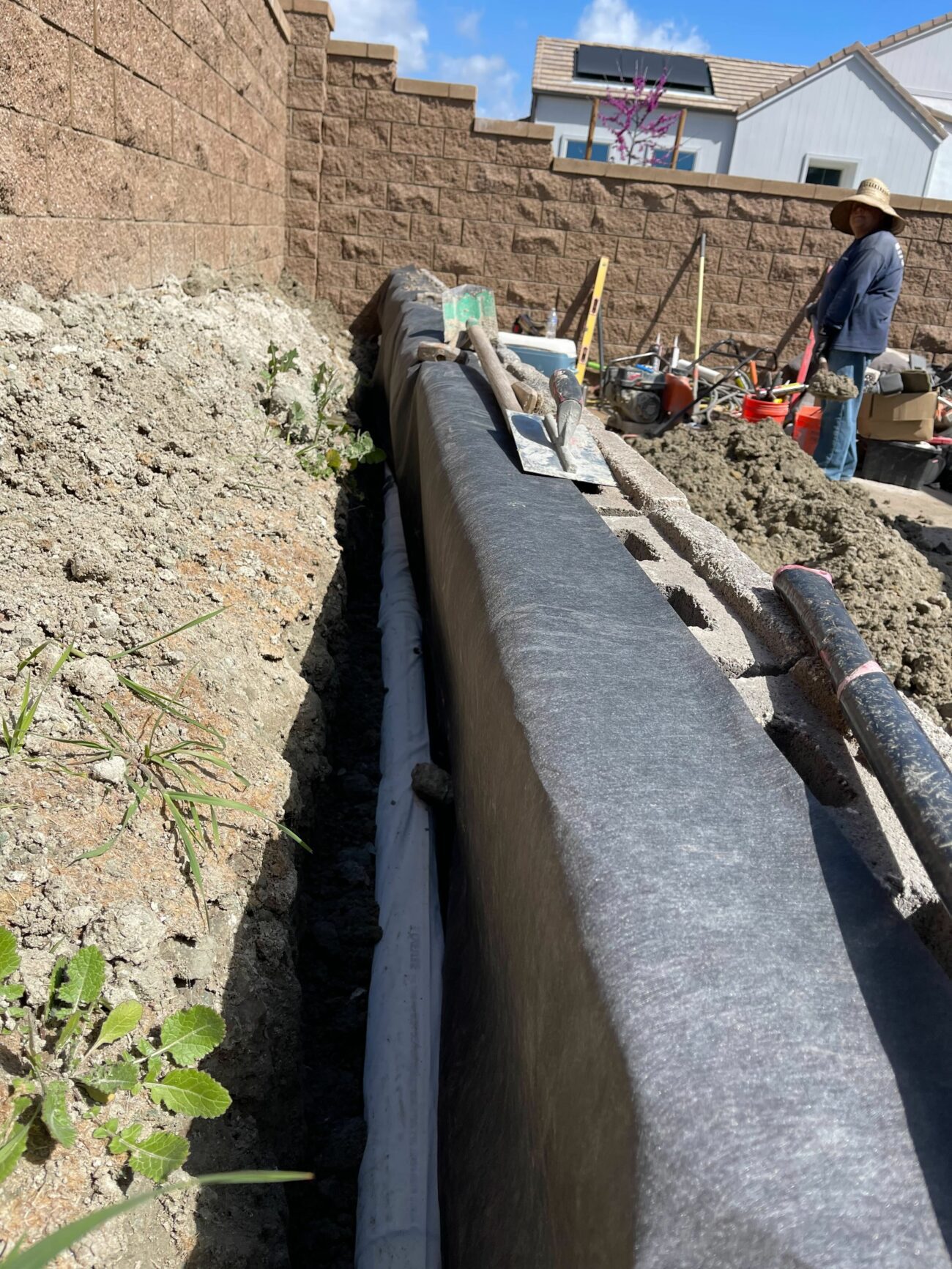 Freshly poured concrete footing in a narrow trench along a block wall, with a trowel resting on top and a worker in backgr...