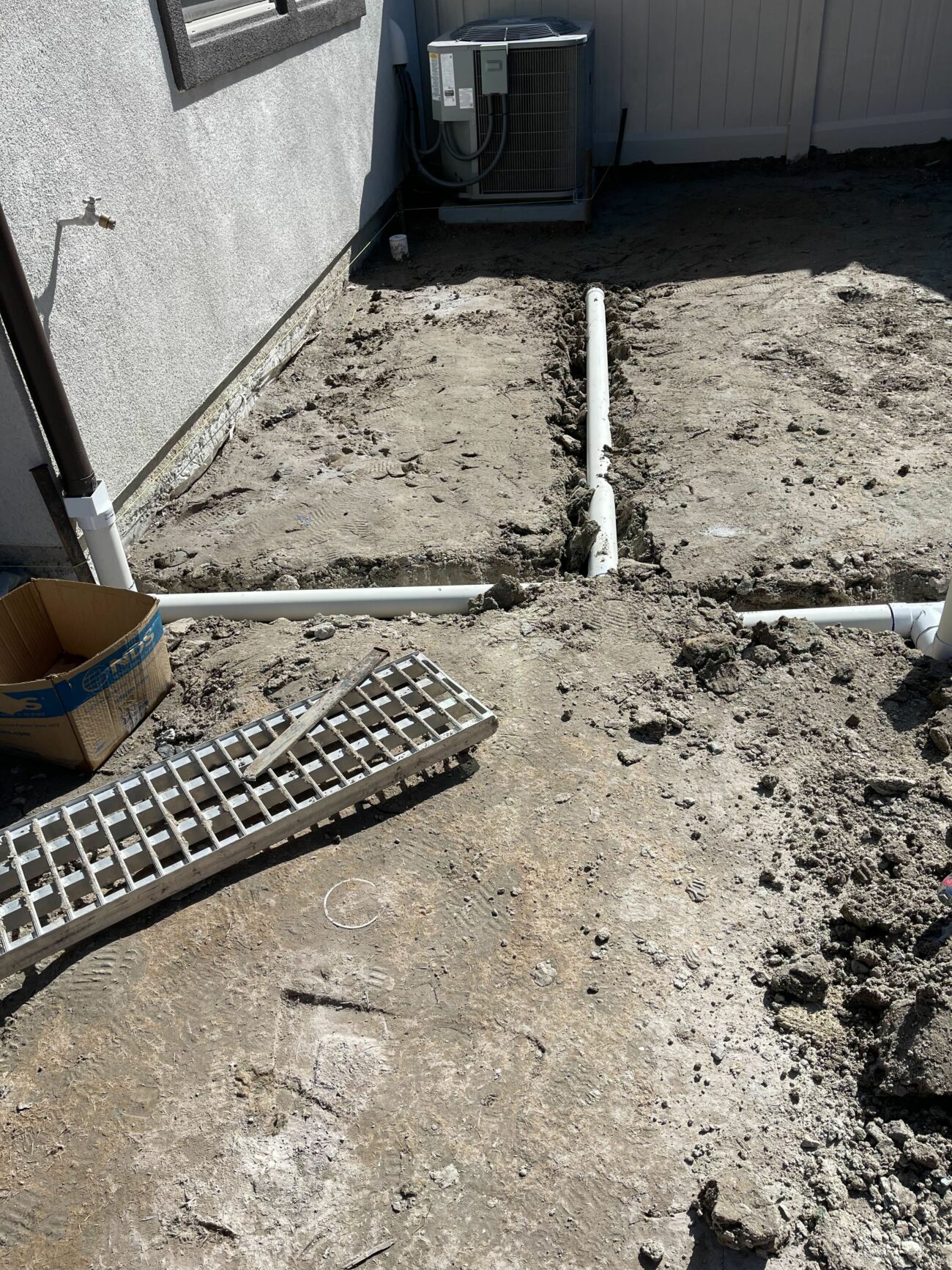Trenched PVC drainage pipes laid in a cross pattern across bare dirt beside a stucco house with an AC unit.