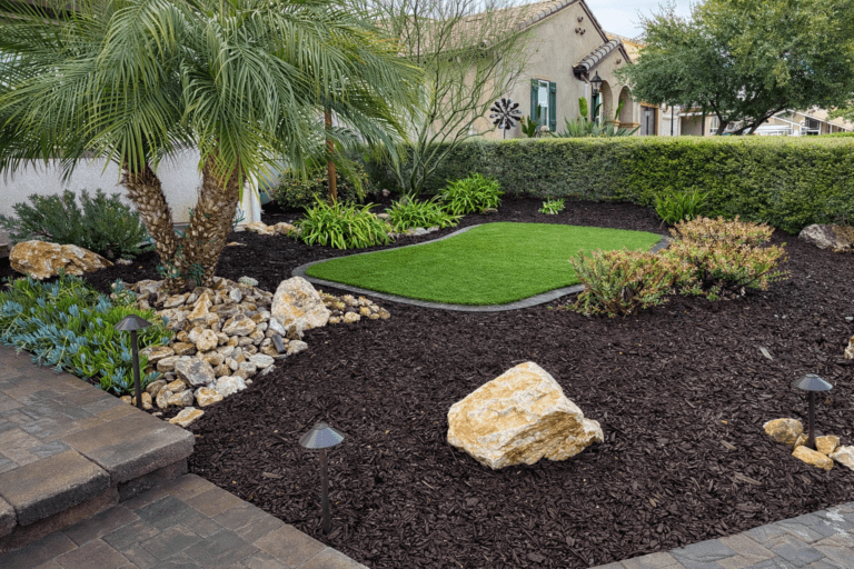 Front yard with dark mulch, boulders, a small palm tree, synthetic turf patch, and low landscape lighting near a paver edge.